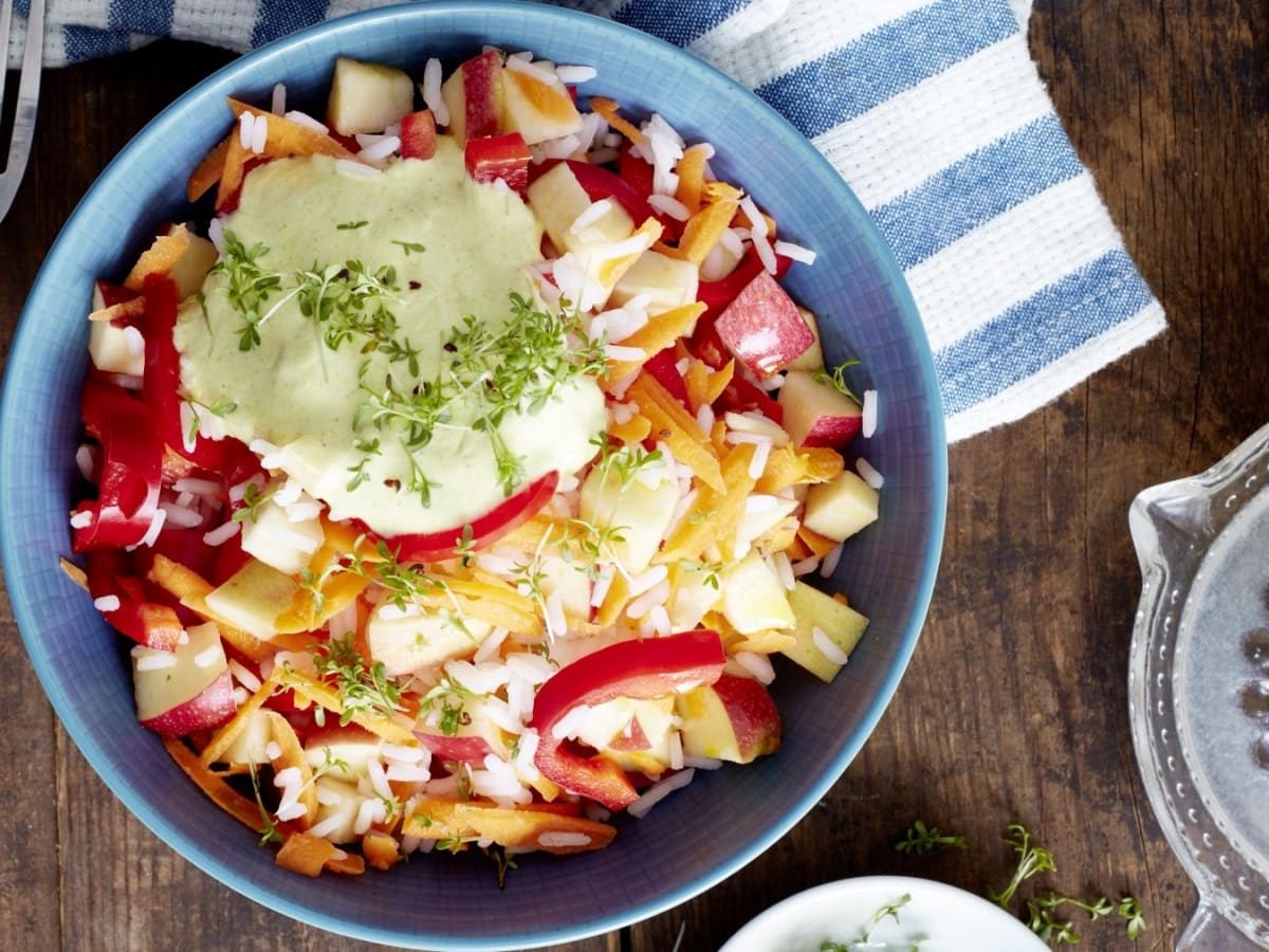 Draufsicht: Dunkler Holzuntergrund mit einem blauen Teller, einer blau-weiß gestreiften Stoffservierte, einer Gabel mit Holzgriff und einer gläsernen Zitronenpresse. Auf dem Teller ein Reissalat mit Apfel, Paprika und einer Sauce aus Joghurt mit frischer Kresse garniert.