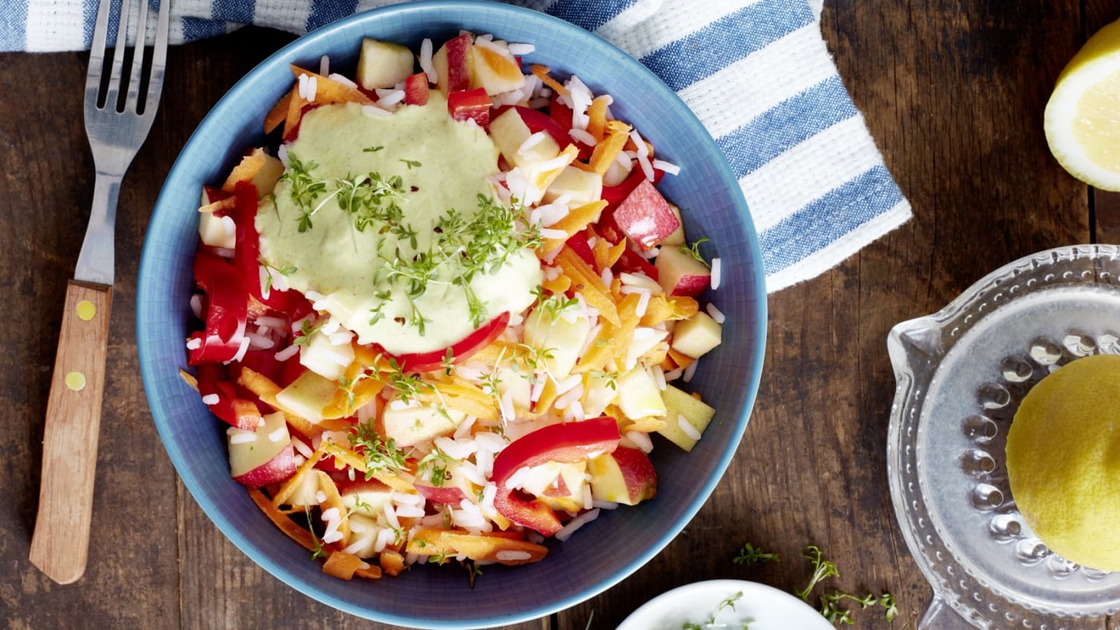 Draufsicht: Dunkler Holzuntergrund mit einem blauen Teller, einer blau-weiß gestreiften Stoffservierte, einer Gabel mit Holzgriff und einer gläsernen Zitronenpresse. Auf dem Teller ein Reissalat mit Apfel, Paprika und einer Sauce aus Joghurt mit frischer Kresse garniert.