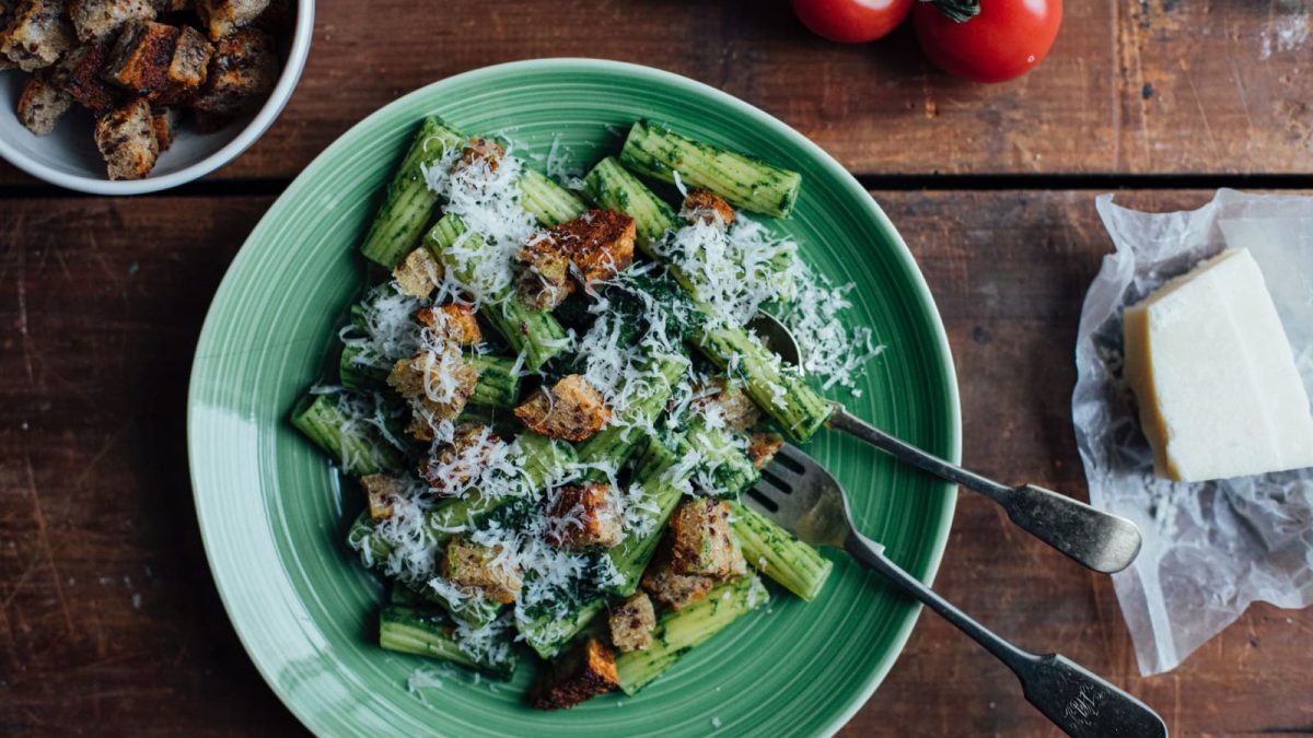 Draufsicht: Hölzerner Tisch auf dem ein grüner Teller mit Rigatoni in einem grünen Pesto steht. Garniert mit Croutons und frisch geriebenem Parmesan. Ebenfalls auf dem Tisch eine Schüssel mit Croutons, frische Tomaten und ein Stück Parmesan.