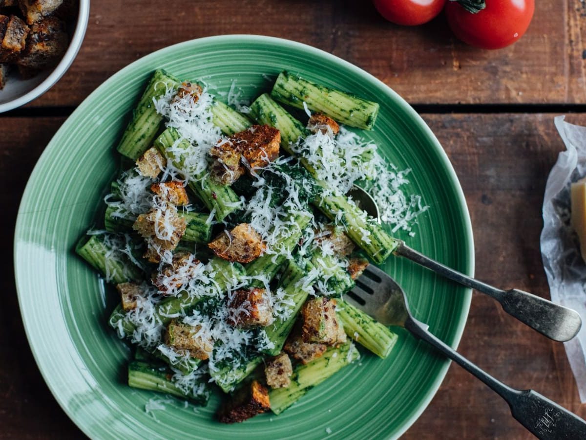 Draufsicht: Hölzerner Tisch auf dem ein grüner Teller mit Rigatoni in einem grünen Pesto steht. Garniert mit Croutons und frisch geriebenem Parmesan. Ebenfalls auf dem Tisch eine Schüssel mit Croutons, frische Tomaten und ein Stück Parmesan.