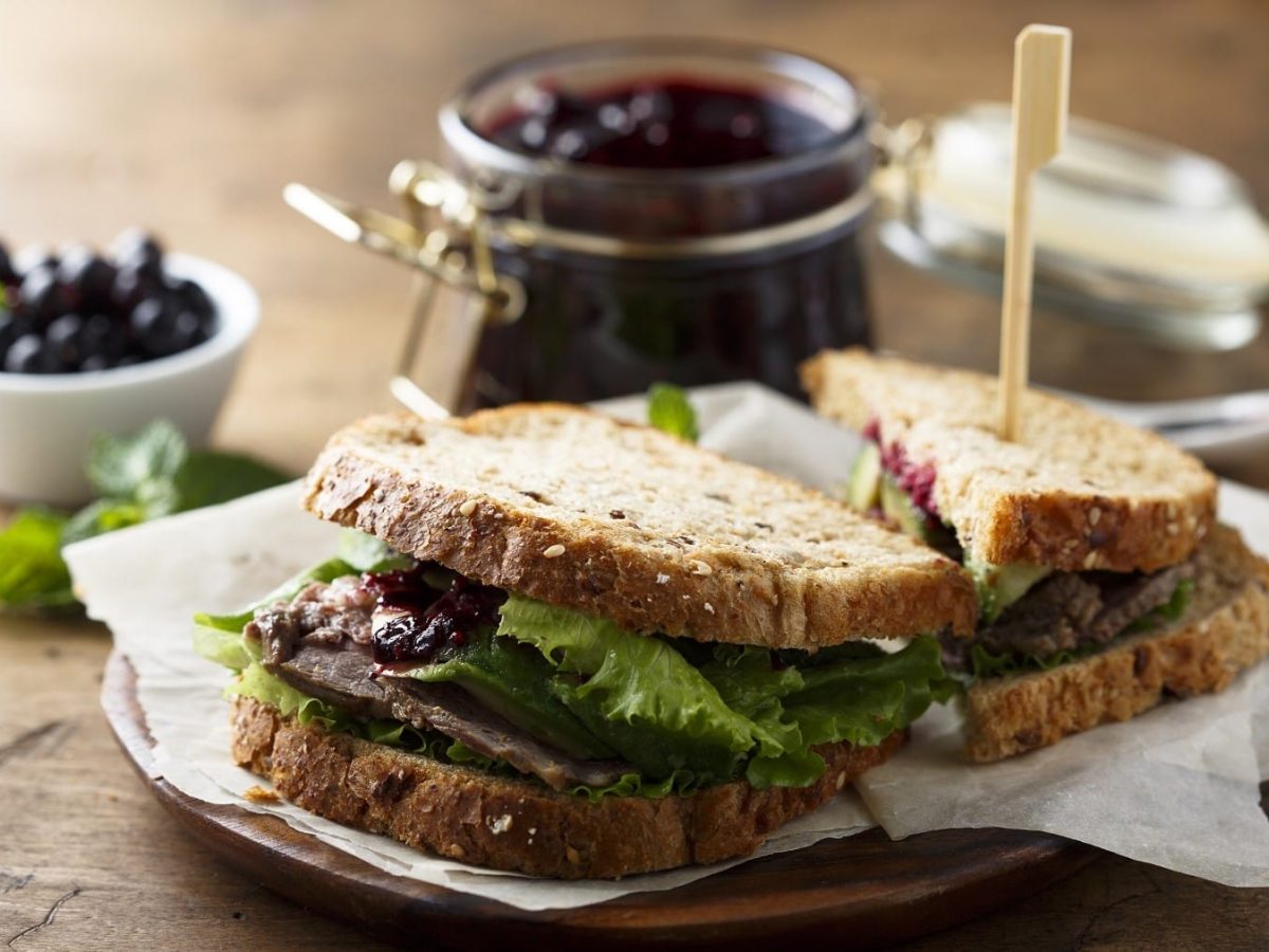 Roastbeef-Sandwich mit Heidebeermarmelade und knackigem Salat, aufgeschnitten serviert auf einem Teller mit noch mehr Marmelade auf einem Holztisch.