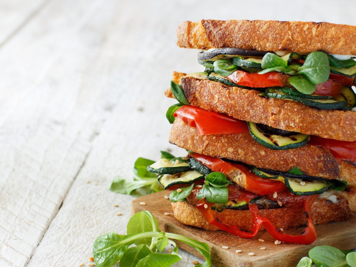 Ein gestapeltes Sandwich mit Grillgemüse, Feldsalat und Tomaten auf hellem Holz.