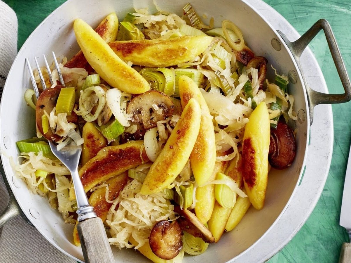 Draufsicht: Grüner hölzerner Untergrund auf dem eine kleine Metallpfanne gefüllt mit gebratenen Schupfnudeln, Sauerkraut, Champignons und frischen Frühlingszwiebeln.