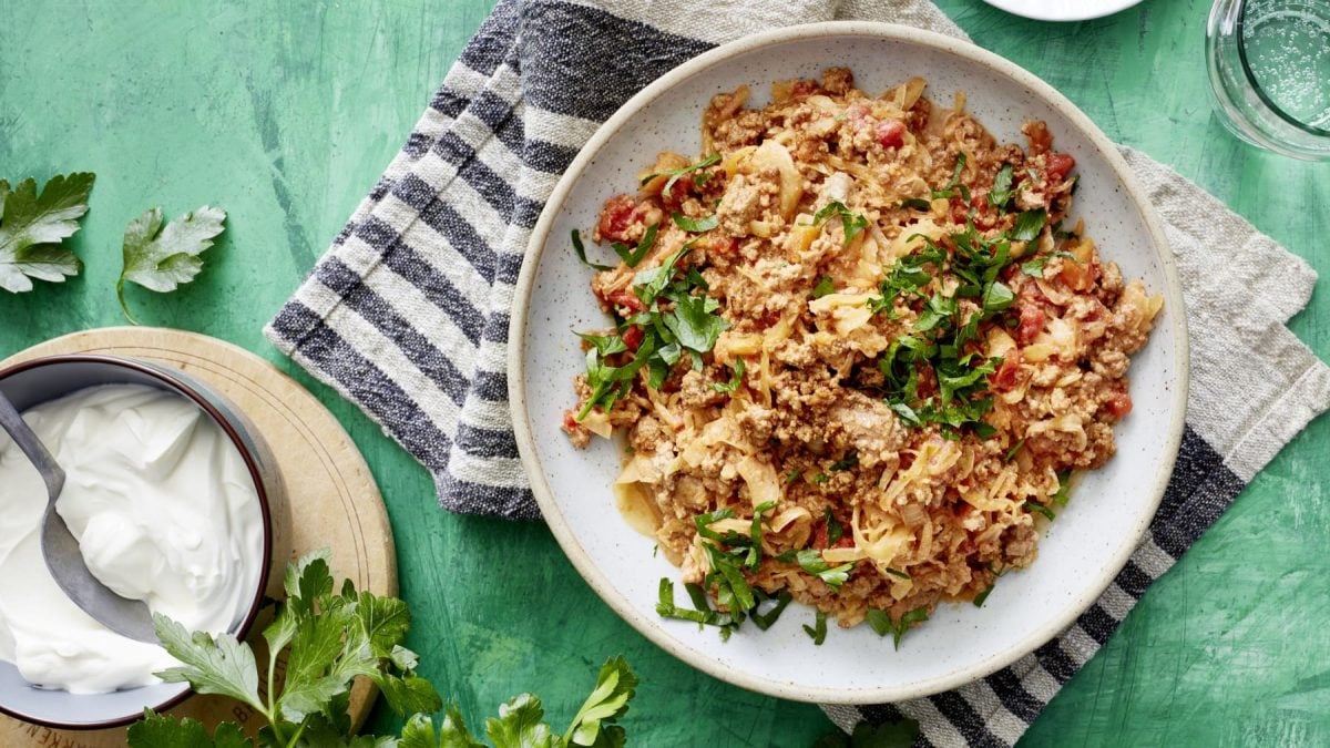 Sauerkraut mit Hack und Tomate neben einer Schüssel saurer Sahne und Petersilie vor grünem Hintergrund.