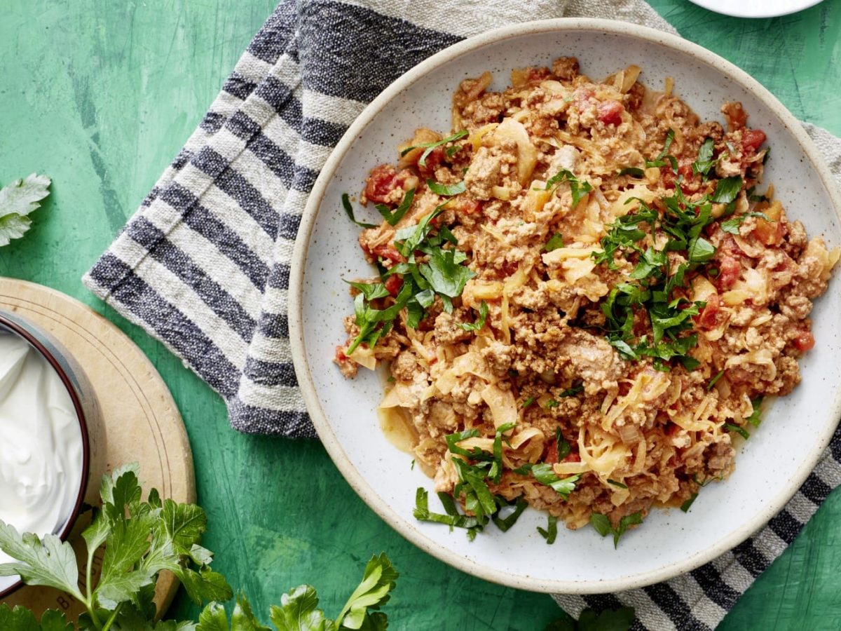 Sauerkraut mit Hack und Tomate neben einer Schüssel saurer Sahne und Petersilie vor grünem Hintergrund.