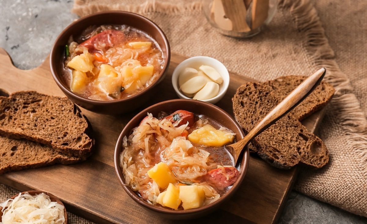 Draufsicht: Zwei dunkle Schalen mit Sauerkrautsuppe und kallser stehen auf einem Holzbrettchen. In einer Suppe ist ein Holzlöffel. Drumherum liegen Brotscheiben, eine helle Schale mit geschältem Knoblauch und ein dunkles Schälchen mit ungeschältem Knoblauchzehen und eine dunkles mit rohem Sauerkraut.