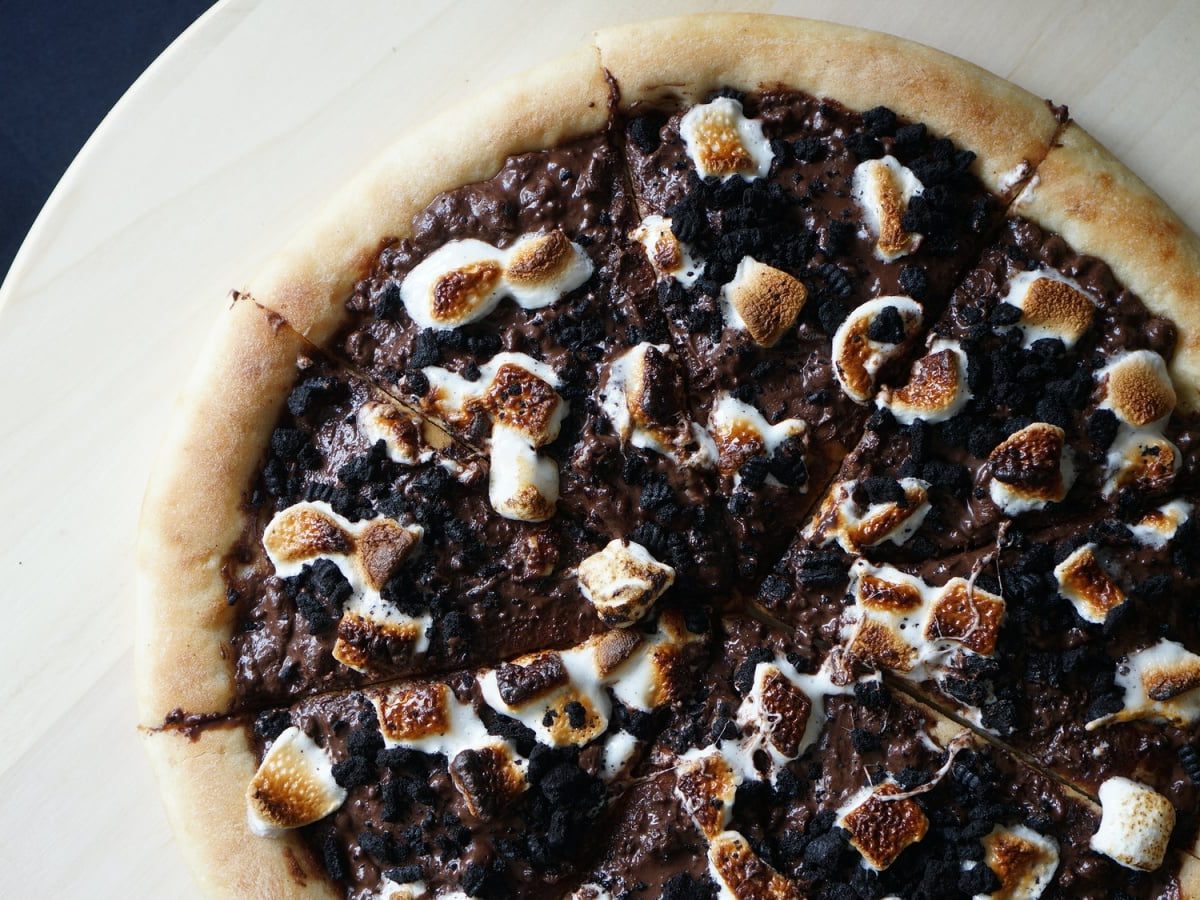 Draufsicht: Dunkler Untergrund mit einem hölzernen Pizzaschieber. Darauf eine Pizza mit Schokoladencreme, Oreo-Stückchen und karamellisierten Marshmallows