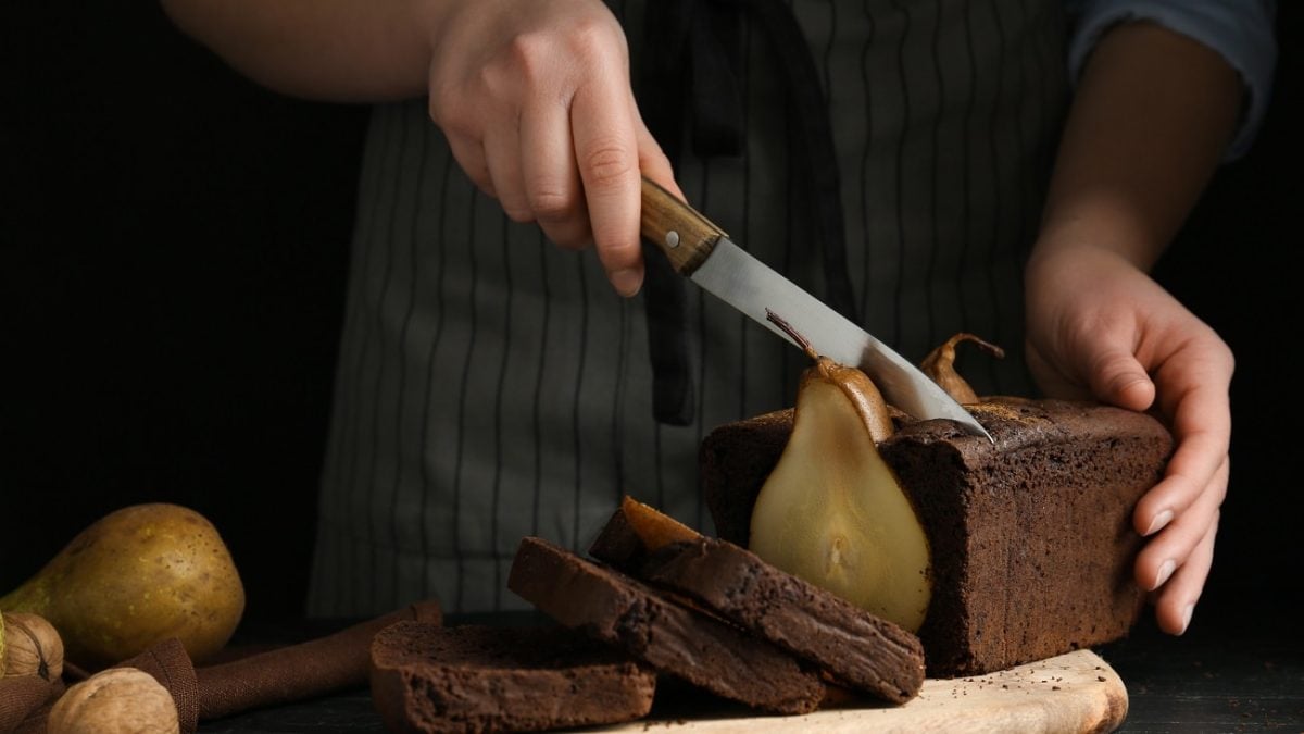 Vor einem dunklen Hintergrund steht ein dunkler Tisch. Auf dem steht ein Schokoladenkuchen mit ganzer Birne. Zwei Hände halten ein großes Küchenmesser mit einer langen Klinge. Diese schneiden den Kuchen an. Neben dem Kuchen liegen Birnen.