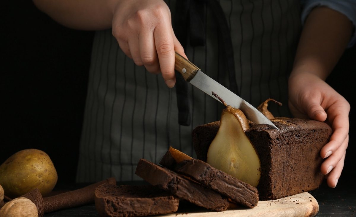 Vor einem dunklen Hintergrund steht ein dunkler Tisch. Auf dem steht ein Schokoladenkuchen mit ganzer Birne. Zwei Hände halten ein großes Küchenmesser mit einer langen Klinge. Diese schneiden den Kuchen an. Neben dem Kuchen liegen Birnen.