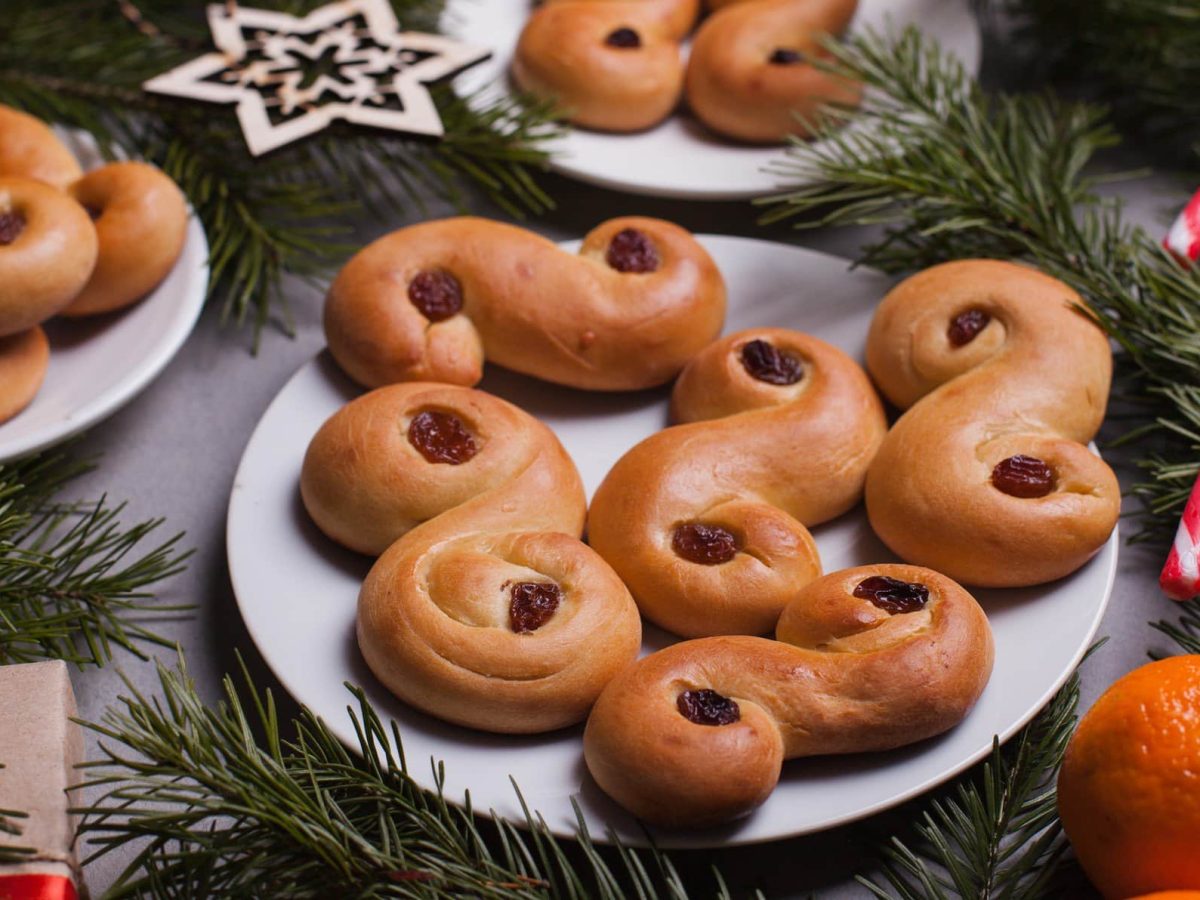 Schwedische Lussekatter auf einem Teller, daneben Tannengrün, Mandarinen und Zuckerstangen.