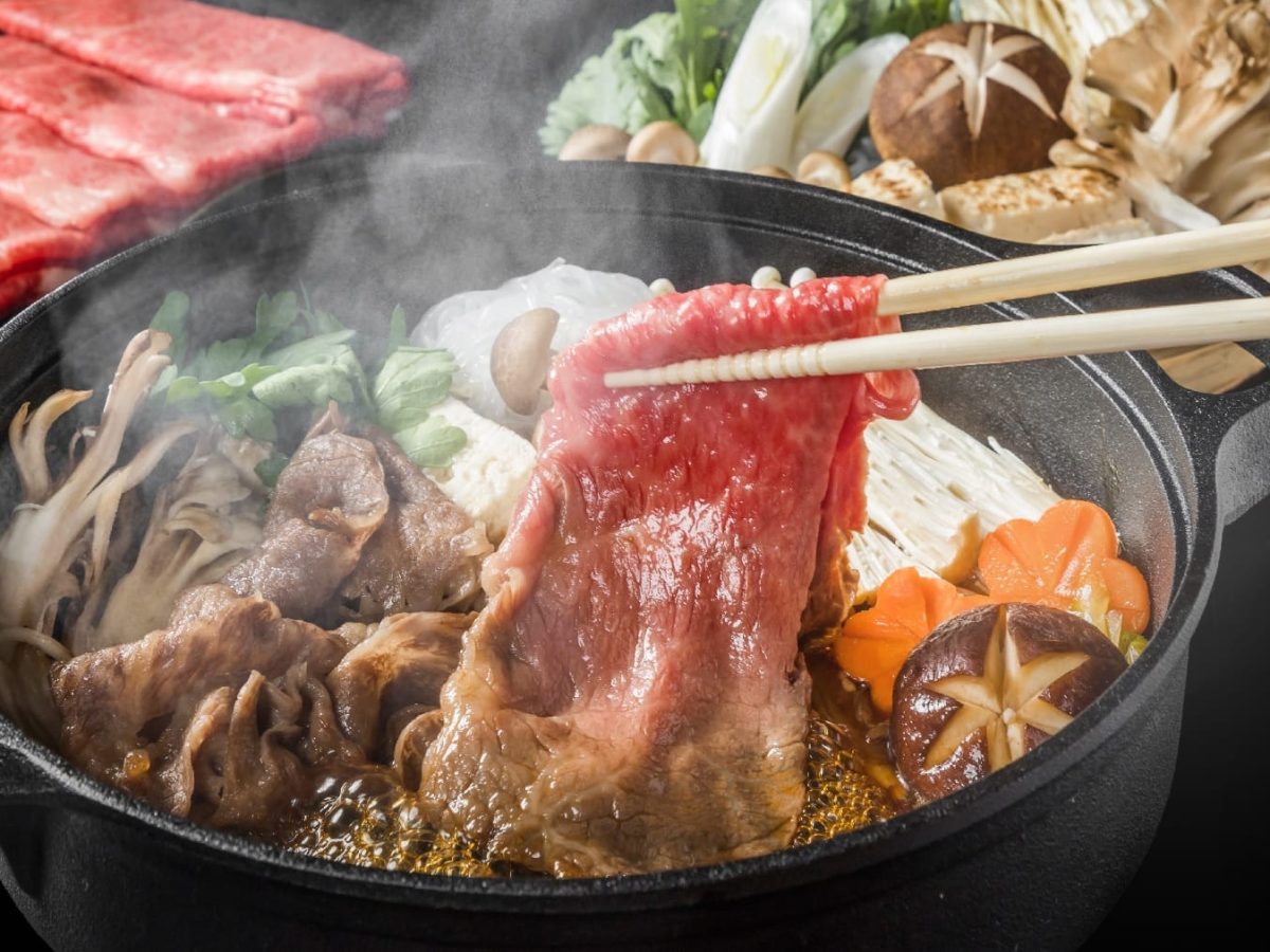 Eine schwarze Schale mit dem Shabu-Shabu. Im Hintergrund viele verschiedene Zutaten und ein schwarzes Brett mit dem Fleisch.