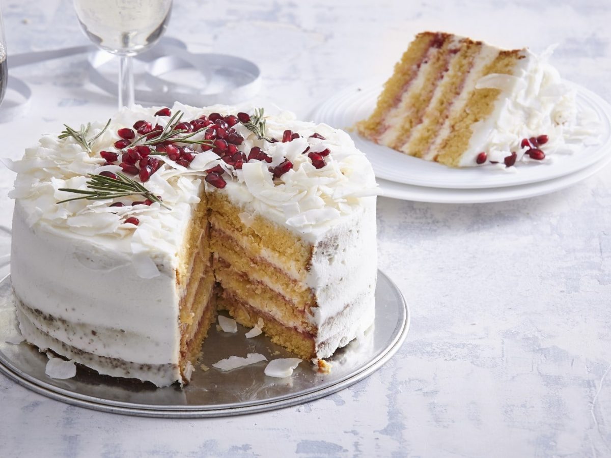 Angeschnittener Silvesterkuchen mit Mandeln und Kokos verziert mit Granatapfelkernen und Rosmarin auf einem Teller vor hellem festlichen Hintergrund.