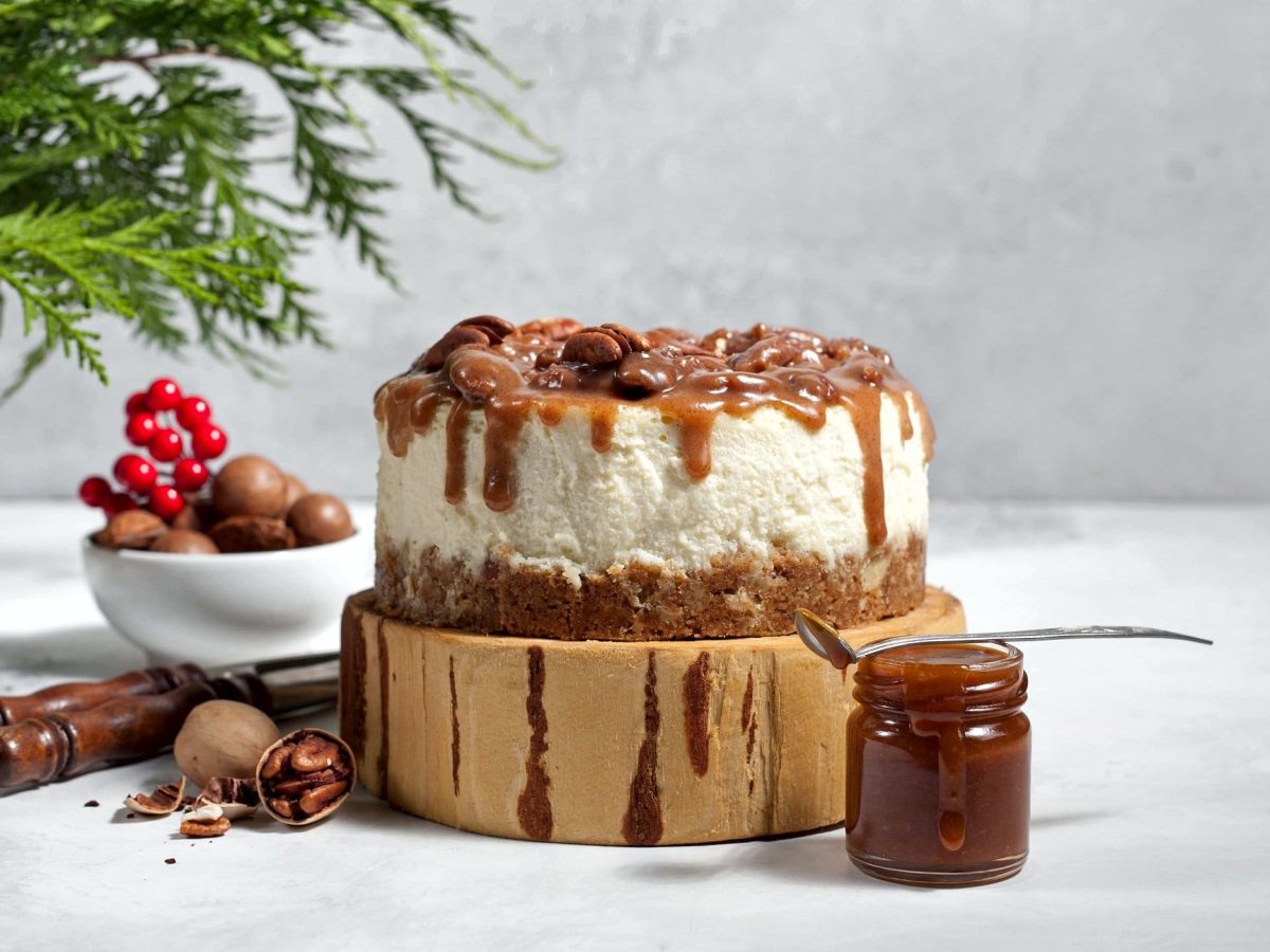 Ein Spekulatius-Cheesecake auf einem Stück Holz, daneben ein Glas mit Karamellsauce und Weihnachtsdeko.