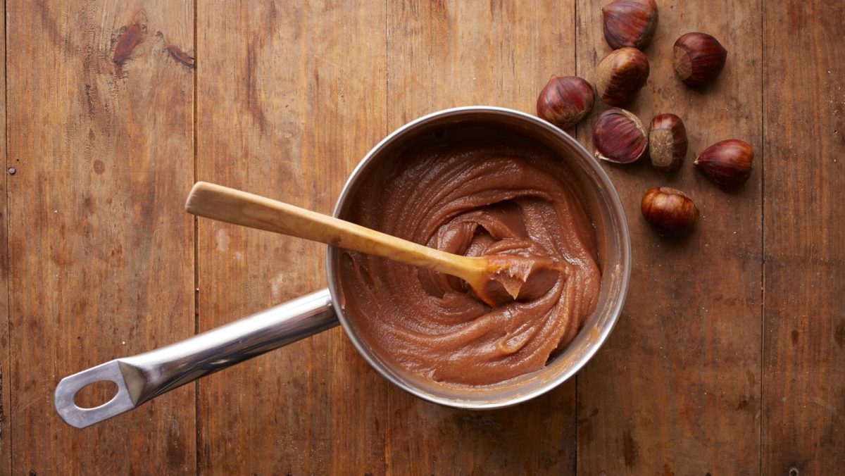 Daraufsicht: Stieltopf auf hölzerner Tischplatte. Gefüllt mit einer braunen süßen Maronencreme. Ein Holzlöffel steckt in der Creme. Dekorativ liegen acht Maronen daneben.