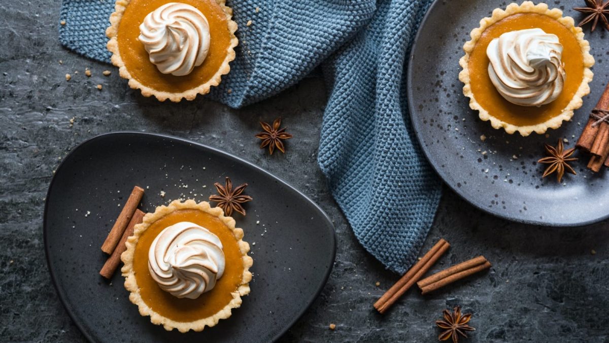 Draufsicht: Dunkler, blau-grauer Untergrund mit zwei Tellern. Drei kleine Sweet Potato Pies mit Baiserhaube. Garniert mit Zimtstangen und echtem Sternanis