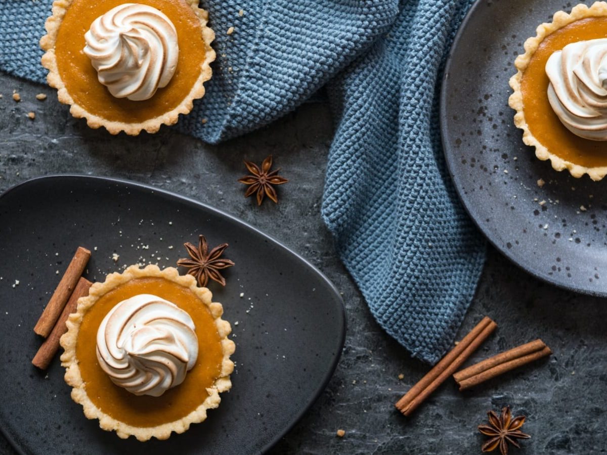 Draufsicht: Dunkler, blau-grauer Untergrund mit zwei Tellern. Drei kleine Sweet Potato Pies mit Baiserhaube. Garniert mit Zimtstangen und echtem Sternanis