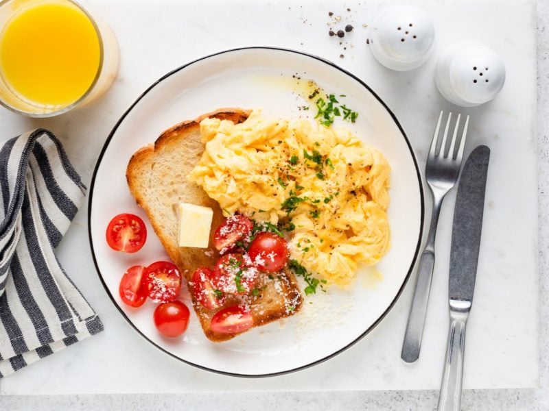 Ein köstliches Rührei auf weißem Grund. Aufnahme aus der Vogelperspektive. Drumherum weitere Frühstücks-Zutaten wie Saft und Tomaten.