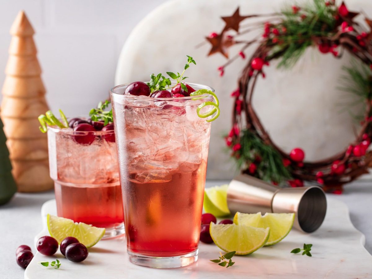 Zwei Gläser Tipsy Rudolph, ein roter Cocktail mit Wodka, darin Oregano, Cranberrys und Eiswürfel. Drumherum Weihnachtsdeko.