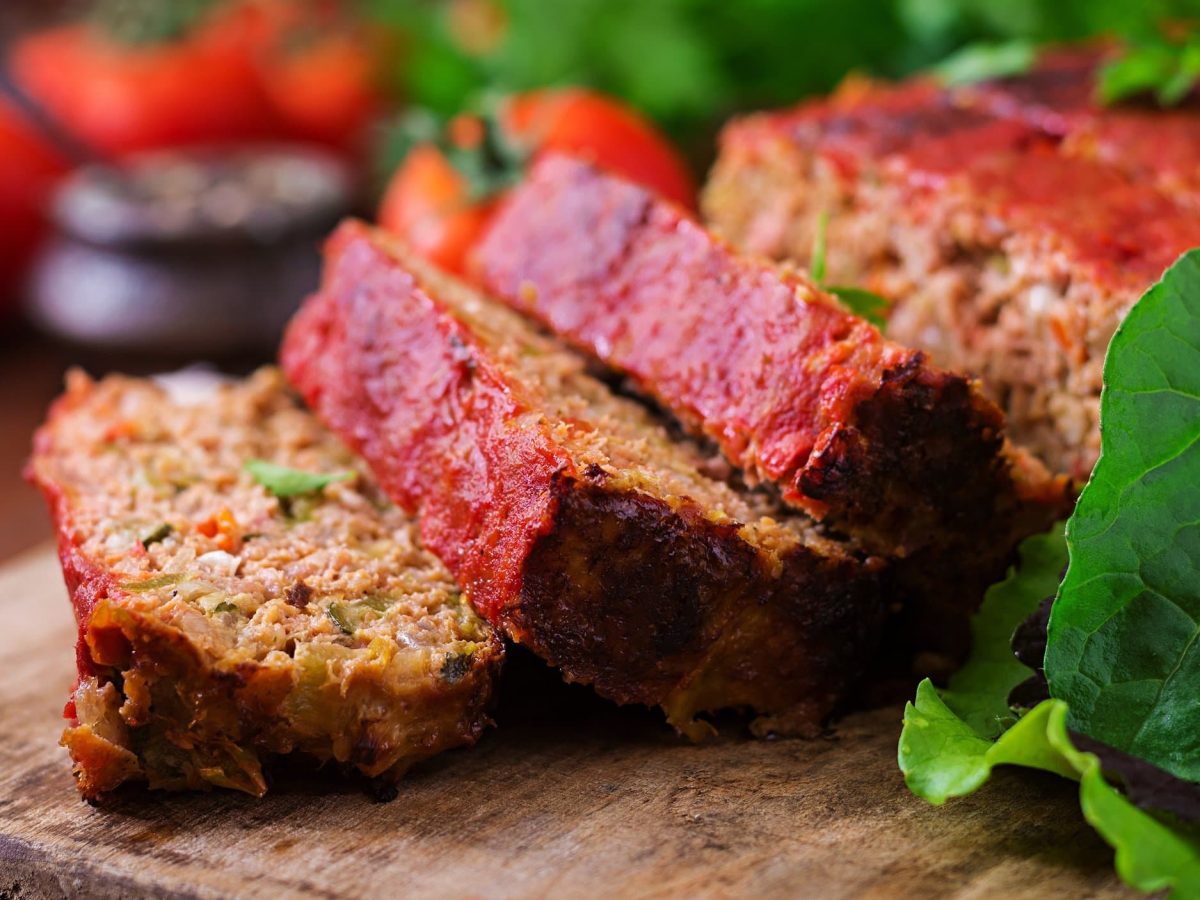 Ein angeschnittener veganer Hackbraten auf einem Brett, daneben frische Tomaten und Salatblätter.