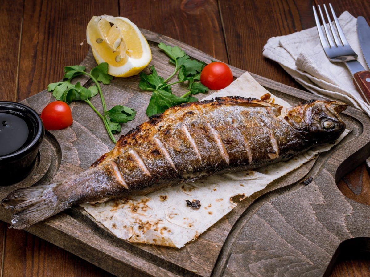 Was ist guter Fisch? Ganze gegrillte Forelle mit Petersilie, Tomaten und Zitrone auf Holzbrett auf Holztisch. Daneben Besteck auf Stoffserviette. Draufsicht.