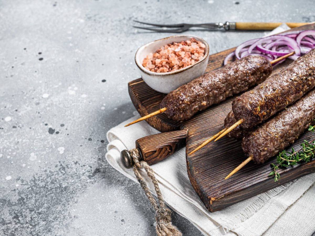 Drei Wild-Köfte-Spieße auf einem Holzbrett mit Thymian und Salz auf grauem Untergrund.