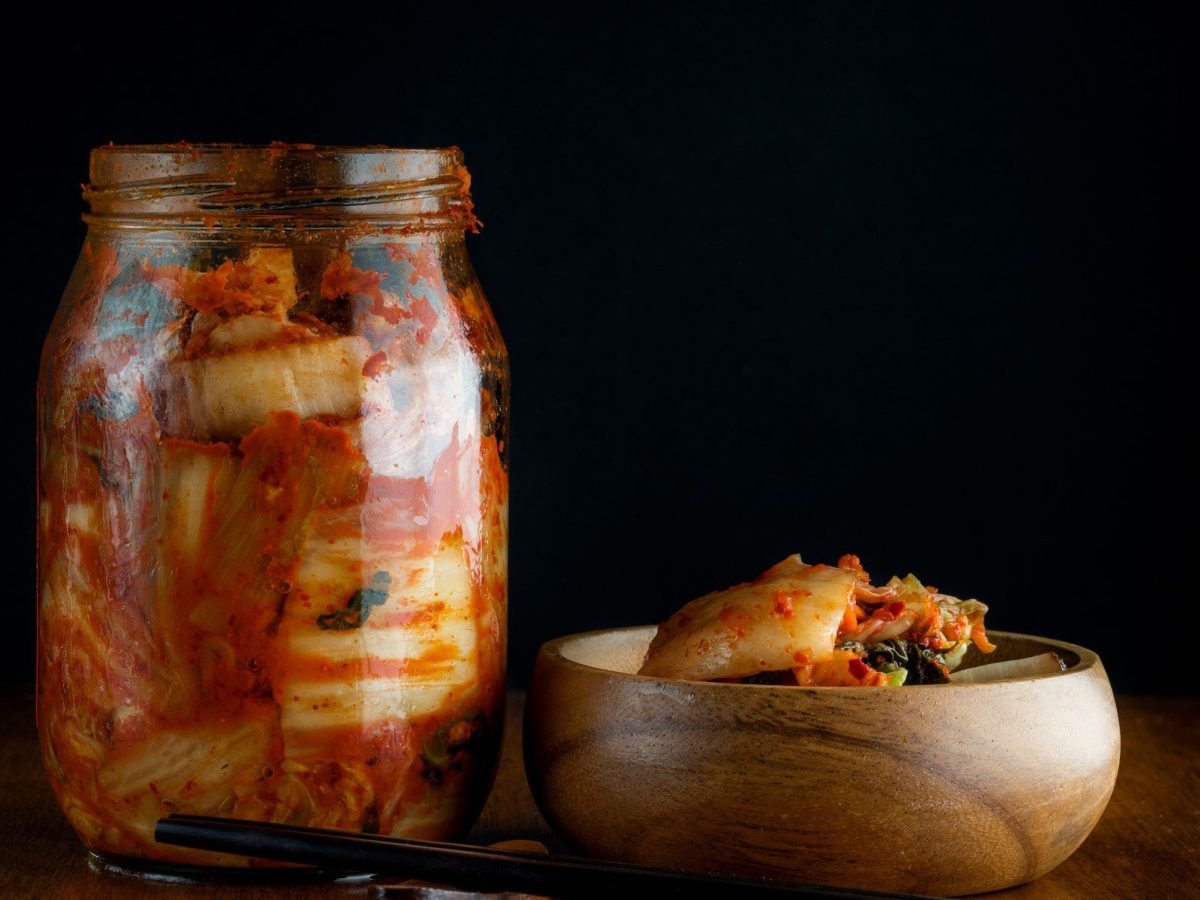 Wirsing haltbar machen mit Wirsing-Kimchi im Glas. Daneben Holzschüssel mit Wirsing-Kimchi und Stäbchen. Frontalansicht.