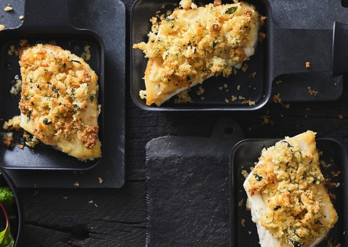 Wolfsbarsch mit Oregano-Parmesan-Kruste aus dem Raclette in drei Pfännchen auf schwarzem Untergrund. Draufsicht.