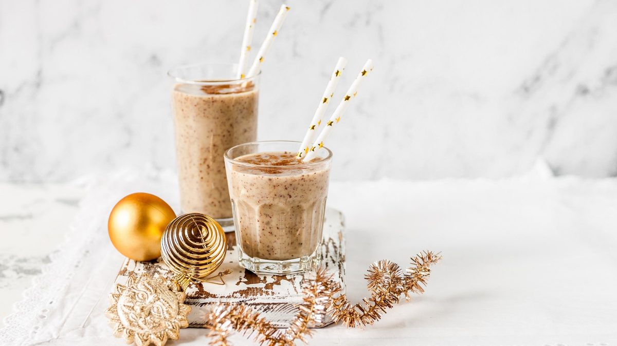 Zwei unterschiedlich große Gläser Zimtschnecken-Milchshake mit Strohhalmen, drumherum Weihnachtskugeln und -girlanden.