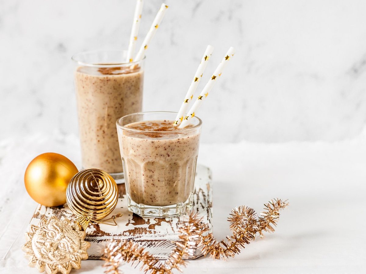 Zwei unterschiedlich große Gläser Zimtschnecken-Milchshake mit Strohhalmen, drumherum Weihnachtskugeln und -girlanden.