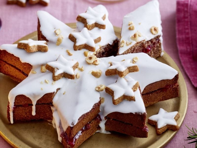 Großer Zimtstern dekoriert mit Zuckerguss, gehackten Nüssen und kleinen Zimtsternen. Serviert auf einem goldenen Küchenteller auf rosa Untergrund.