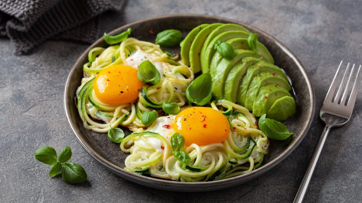 Ein schwarzer Teller auf grauem Untergrund mit zwei Spiegeleiern. Zucchini-Nudeln und einer geschnittenen Avocado. Alles garniert mit Basilikumblättern.