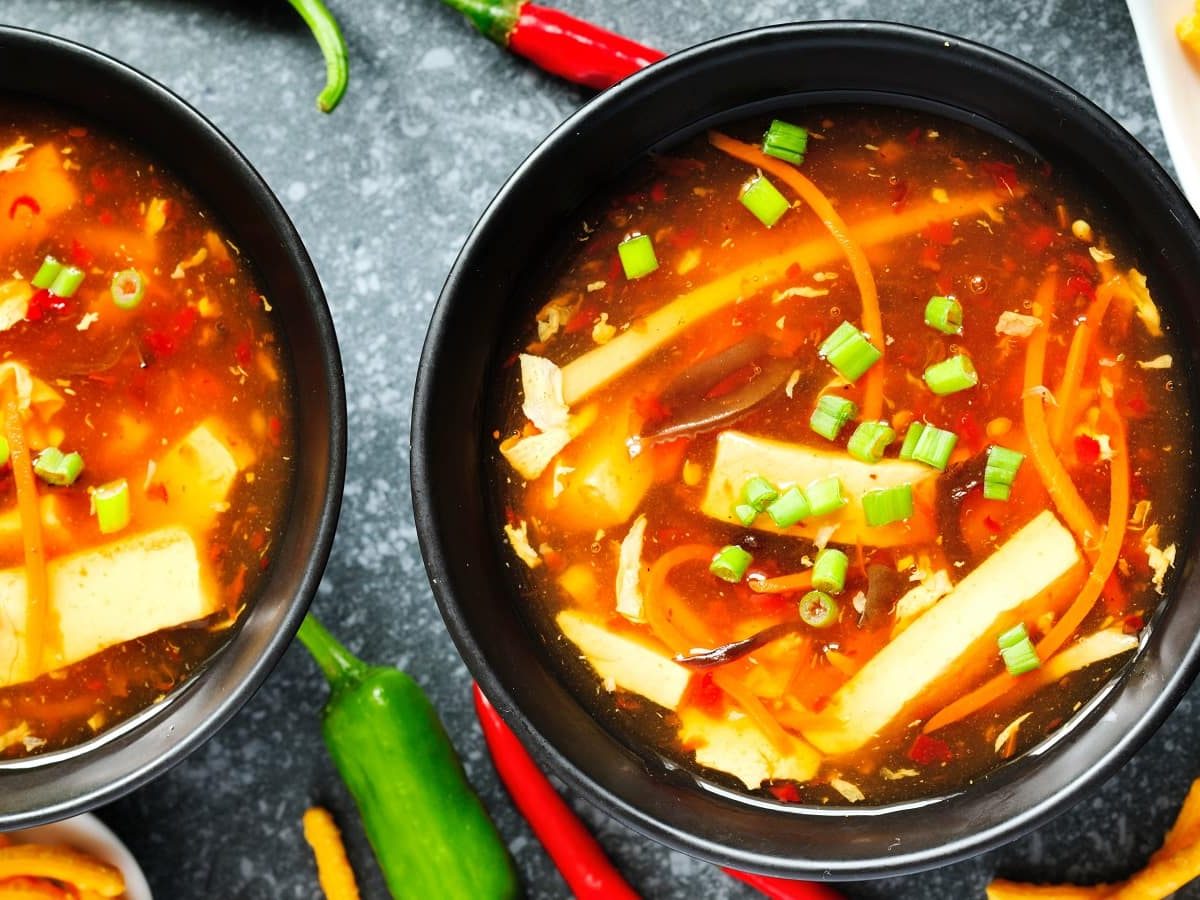 Zwei schwarze Schüsseln mit Sauer-Scharf-Suppe neben frischen Chilis.