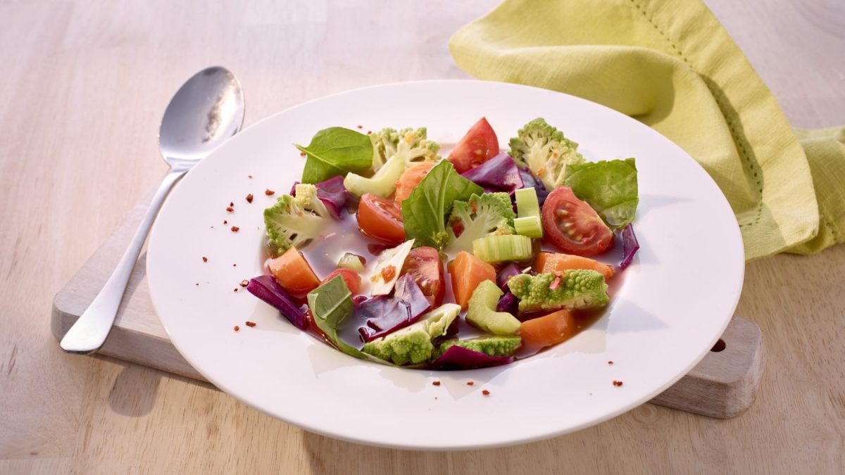 Detox-Suppe mit Tomaten und Staudensellerie, Rote Bete, frischem Basilikum und Kohl. Serviert in einer großen, weißen Suppenschüssel auf einem Holztisch.