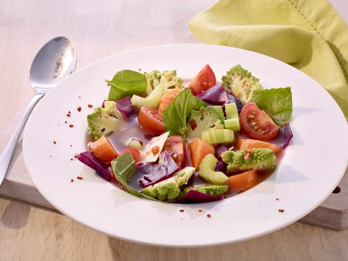 Detox-Suppe mit Tomaten und Staudensellerie, Rote Bete, frischem Basilikum und Kohl. Serviert in einer großen, weißen Suppenschüssel auf einem Holztisch.