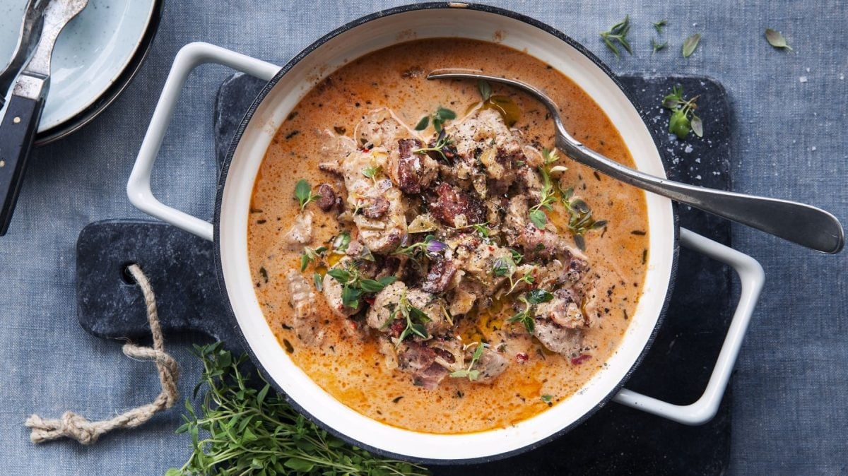 In einem Topf ist ein Eintopf mit Schweinefilet, Speck und getrockneten Tomaten angerichtet. Er ist mit Thymian garniert.