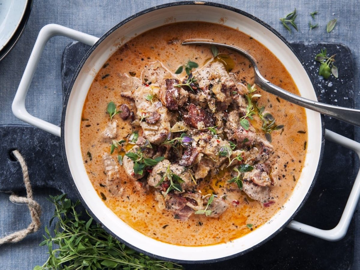 In einem Topf ist ein Eintopf mit Schweinefilet, Speck und getrockneten Tomaten angerichtet. Er ist mit Thymian garniert.