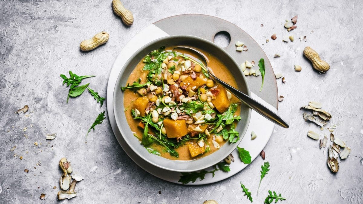 Kürbissuppe mit Erdnüssen, garniert mit frischem Rucola und gerösteten Erdnüssen, serviert in einer weißen Schale auf steinernem Untergrund.