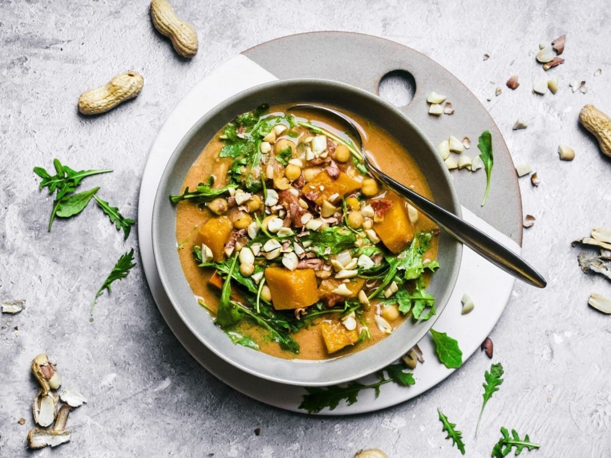 Kürbissuppe mit Erdnüssen, garniert mit frischem Rucola und gerösteten Erdnüssen, serviert in einer weißen Schale auf steinernem Untergrund.