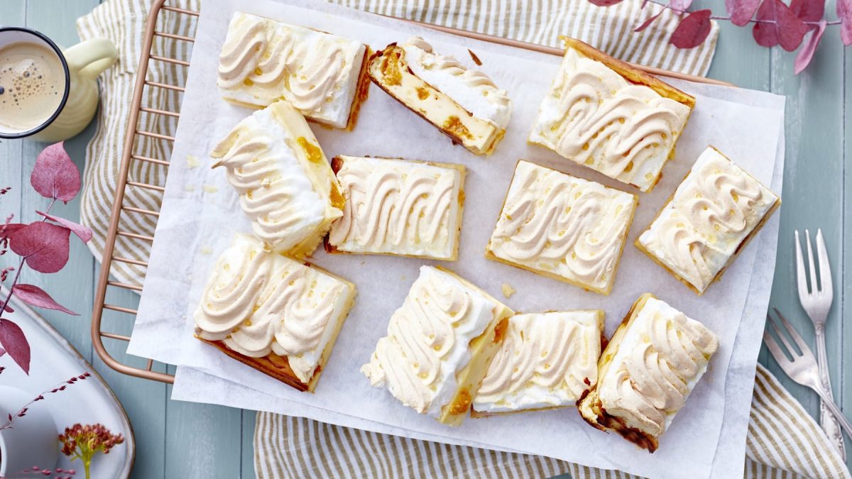 Mandarinen-Schnitten mit Baiser, als Blechkuchen angeschnitten und auf einem weißen Backpapier auf einem hellen Tisch serviert.