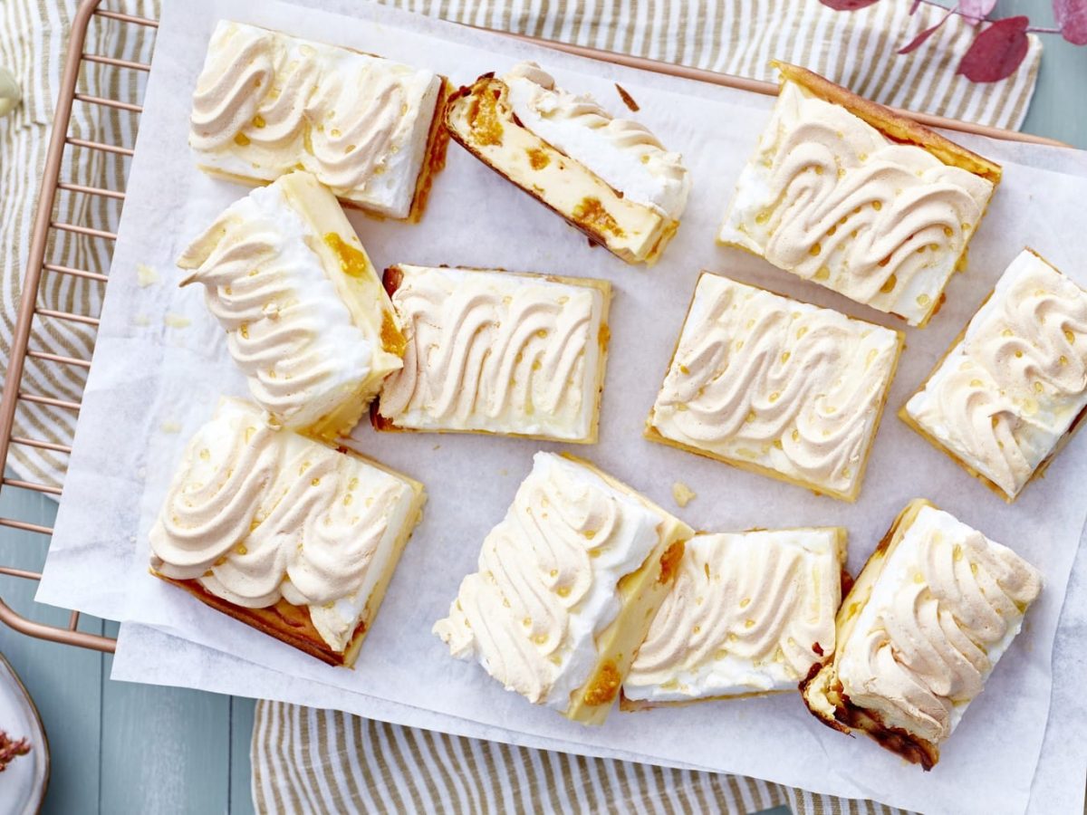 Mandarinen-Schnitten mit Baiser, als Blechkuchen angeschnitten und auf einem weißen Backpapier auf einem hellen Tisch serviert.