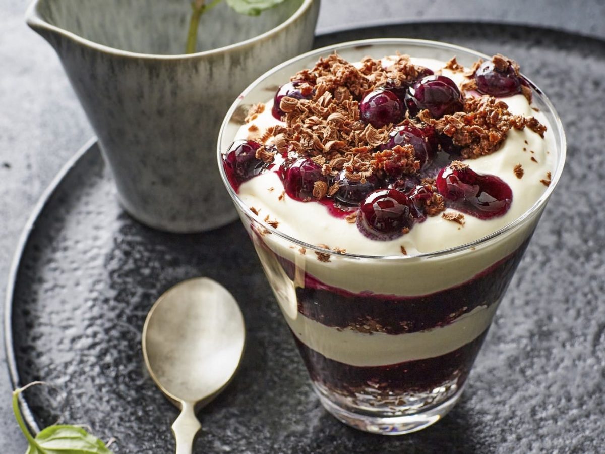 Stippmilch mit Blaubeerkompott und Schokoladenstreuseln, geschichtet mit dunkler Pumpernickelschokolade und serviert in einem durchsichtigen Dessertglas.