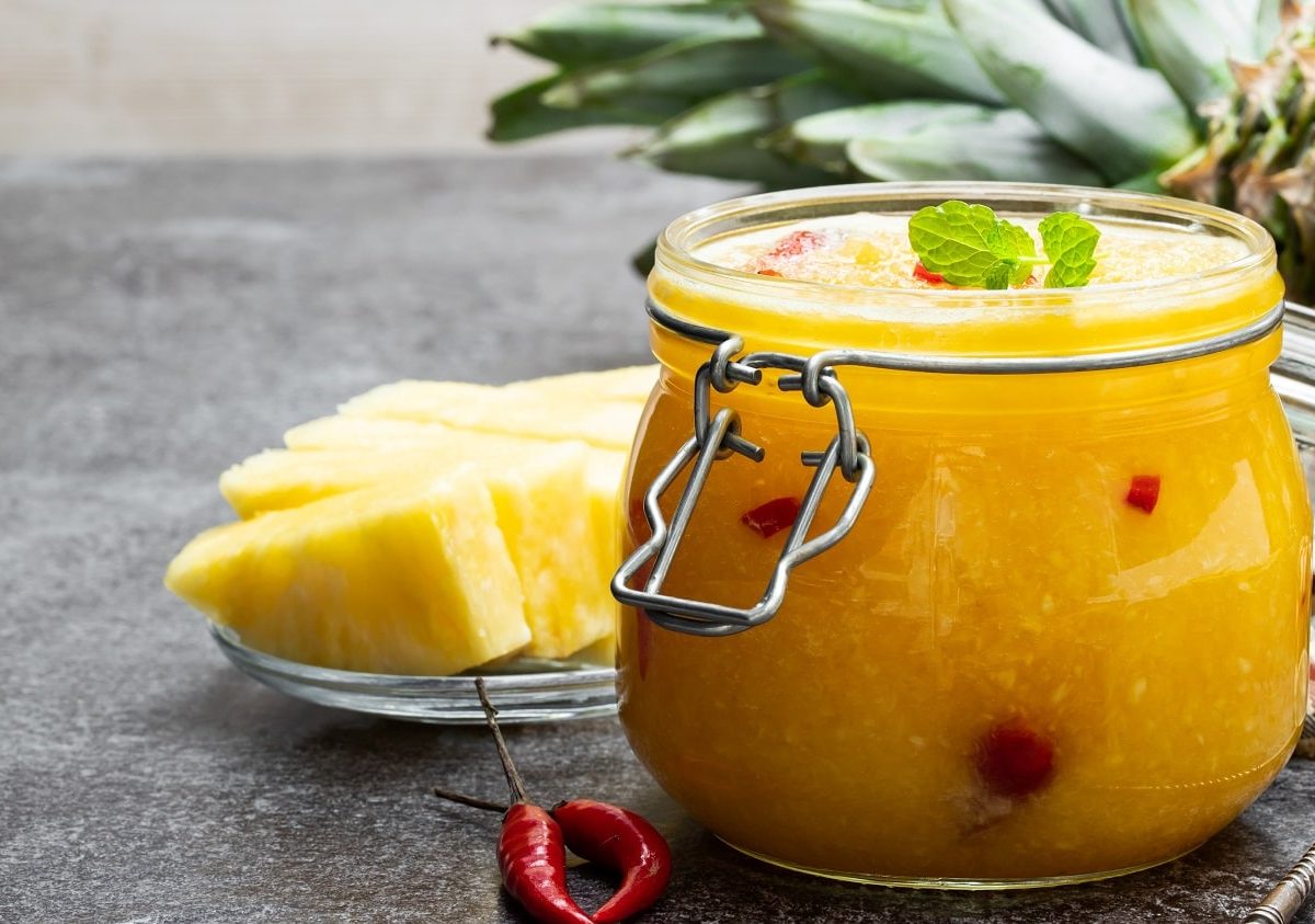 Frontalsicht: Ein Ein Einmachglas Elena Zajchikova steht auf einem Holztisch. Daneben lieben Ananasstücke und Chilis. Im Hintergrund liegt noch eine Ananas.