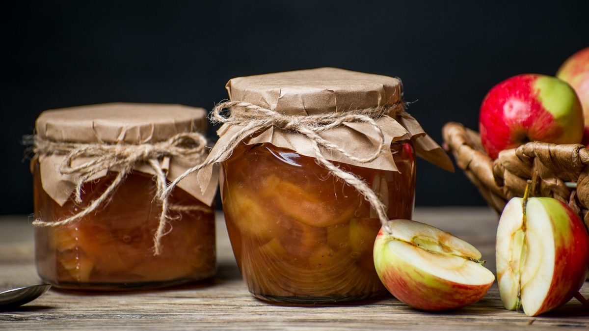 frontalsicht: zwei einweckgläse mit einem apfel-minz-relish stehen auf einem untergrund aus holz. der hintergrund ist dunkel. neben den gläsern, liegern halbierte und ganze äpfel.