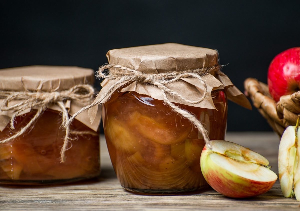 frontalsicht: zwei einweckgläse mit einem apfel-minz-relish stehen auf einem untergrund aus holz. der hintergrund ist dunkel. neben den gläsern, liegern halbierte und ganze äpfel.