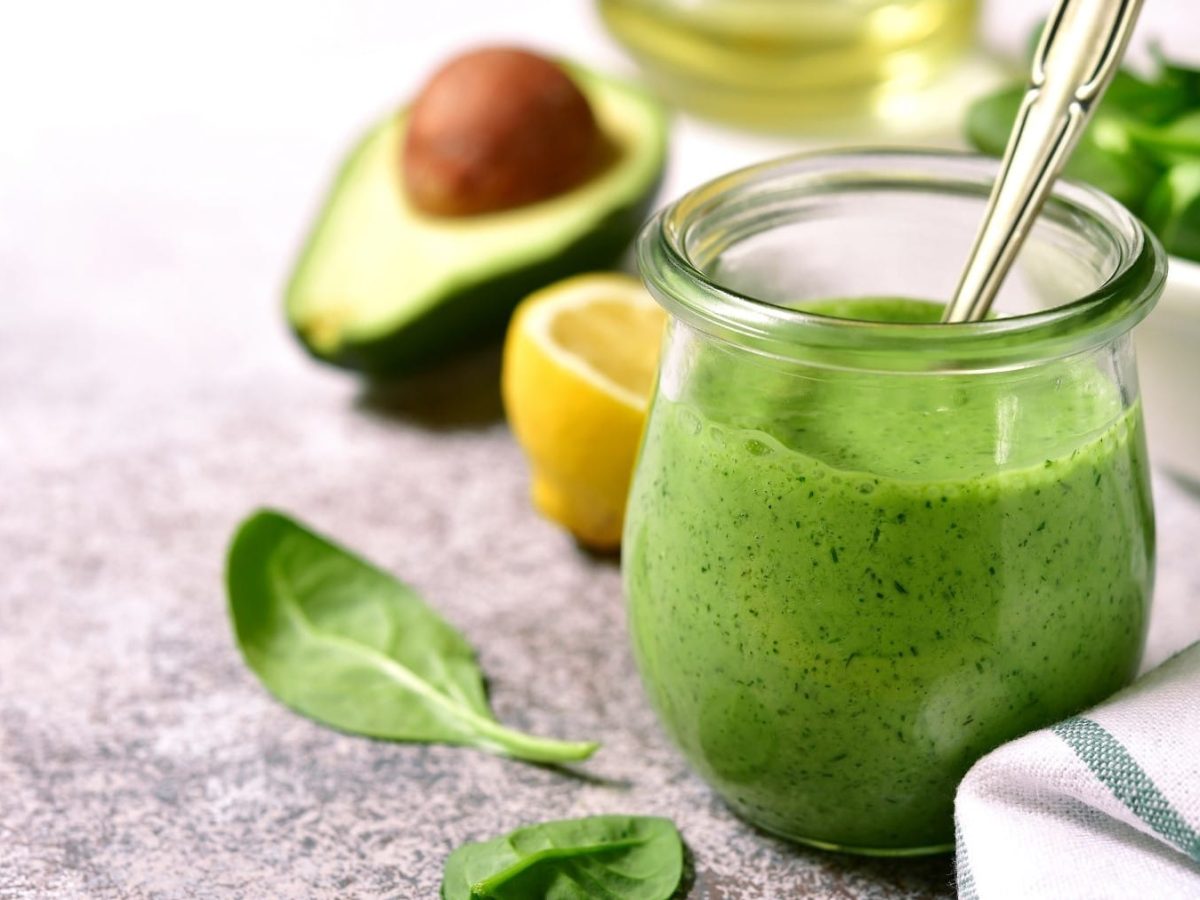 Ein Glas mit dem Avocado-Dressing auf einem grauen Tisch mit Zitrone, Babyspinat und Avocado.