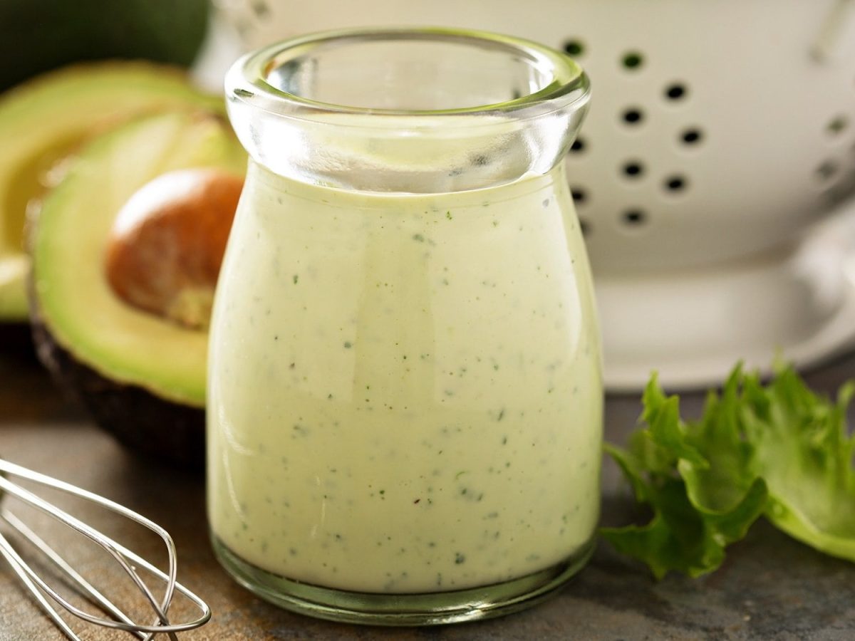 Eine kleine Glaskaraffe mit Avocado Ranch Dressing, dahinter unscharf eine halbierte Avocado, Salatblätter und ein Metallsieb.