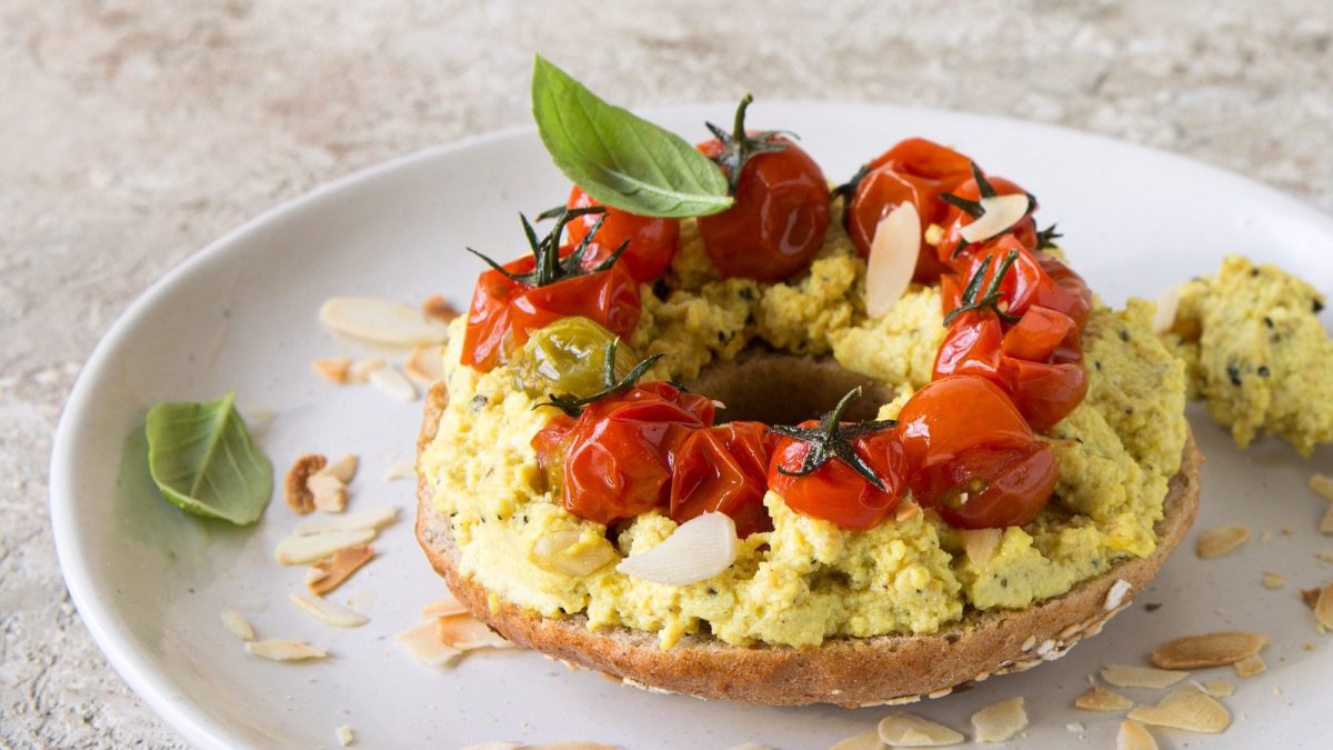 Ein Bagel-Sandwich mit Curry-Tofu, Basilikum, Mandelblättchen und Tomaten auf einem Teller.