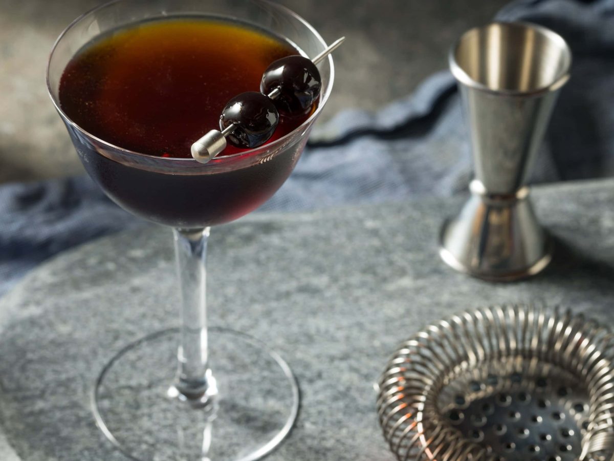 Black Manhattan Cocktail in Cocktailschale auf grauem Holztisch. Daneben Sieb und Barmaß. Draufsicht.