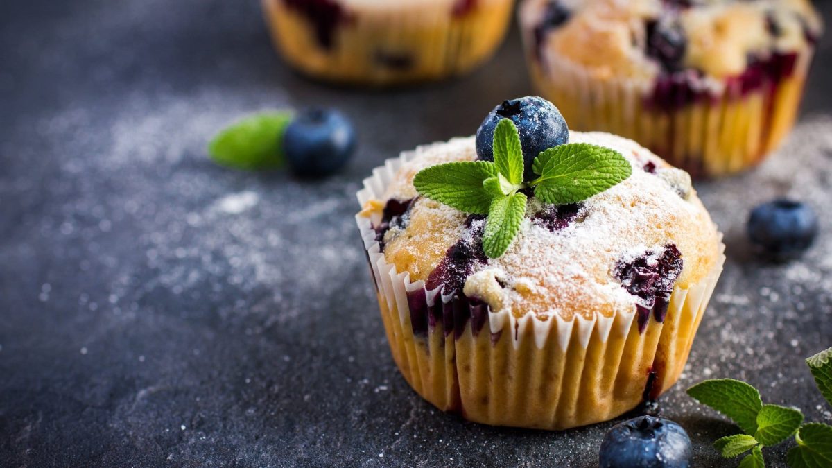 Ein Blaubeer-Zitronen-Muffin mit Puderzucker und Minzblättchen garniert, im Hintergrund unscharf weitere Muffins zu sehen.