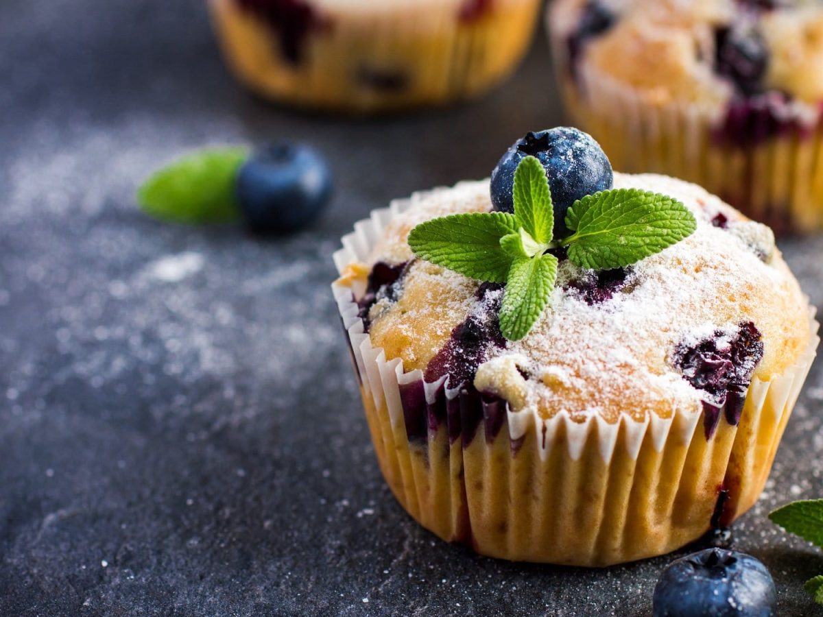 Ein Blaubeer-Zitronen-Muffin mit Puderzucker und Minzblättchen garniert, im Hintergrund unscharf weitere Muffins zu sehen.